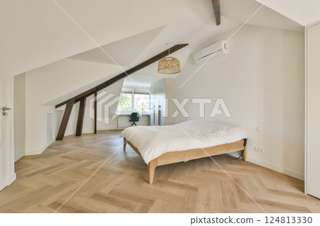 A stylish modern bedroom featuring a wooden floor, slanted ceiling, and a cozy bed. The space is well-lit with natural light from a nearby window. 124813330