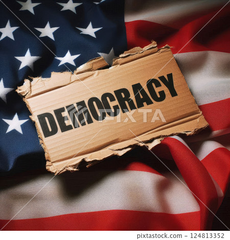 A piece of torn cardboard with the word "democracy" written on it lies on an American flag. Political concept for news sites A piece of torn cardboard with the word "democracy" written on it lies on an American flag. Political concept for news sites 124813352