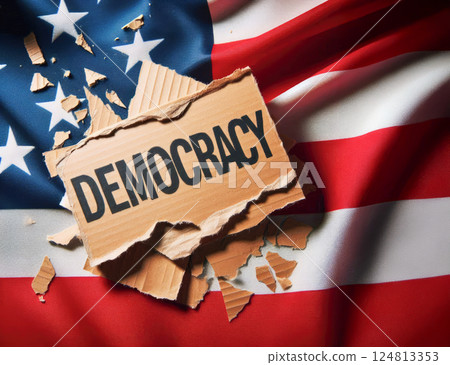 A piece of torn cardboard with the word "democracy" written on it lies on an American flag. Political concept for news sites 124813353