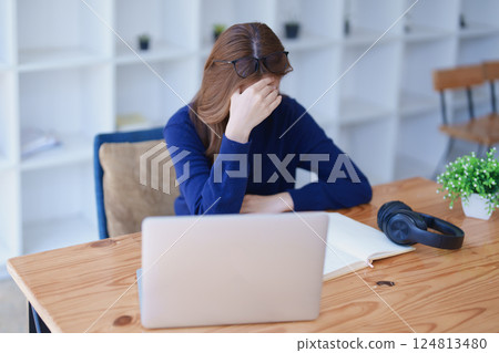 Stressed young woman overwhelmed by e-learning studies at desk with laptop, notebook, headphones, hands clasped to her face in frustration 124813480