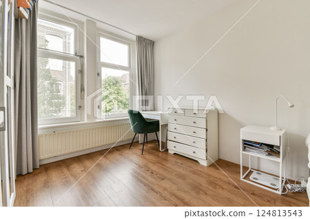 A spacious, minimalist bedroom featuring large windows, natural light, and a simple yet elegant decor with a desk and a comfortable armchair. 124813543