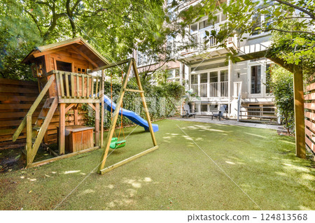 A vibrant playground set in a lush green garden featuring a wooden playhouse, a swing, and a blue slide, perfect for outdoor child play. 124813568