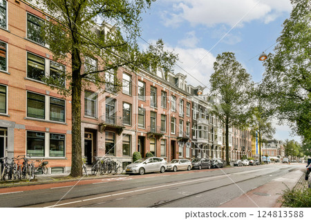 A picturesque urban street scene featuring historic buildings, green trees, parked bicycles, and cars under a clear sky, showcasing a blend of city life and nature. 124813588