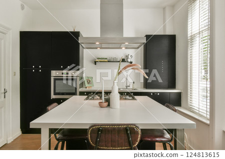 A stylish and contemporary kitchen featuring a sleek design with black cabinetry, a spacious table, and a bright atmosphere enhanced by natural light. 124813615