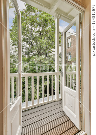 A well-lit balcony opens up to a lush green view, framed by white doors and wooden flooring, inviting nature into the living space. 124813675