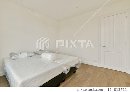 A spacious and modern minimalist bedroom featuring a neatly arranged bed with protective covers, light wooden flooring, and neutral wall colors. A spacious and modern minimalist bedroom featuring a neatly arranged bed with protective covers, light wooden flooring, and neutral wall colors. 124813711