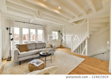 A stylish and contemporary living room featuring a gray sofa, wooden flooring, and bright natural light pouring through large windows, creating a warm atmosphere. 124813919