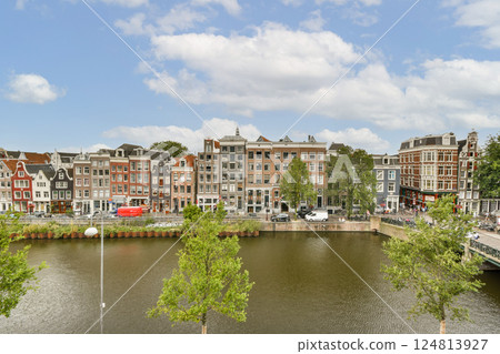 A beautiful view of a riverfront lined with historic buildings and lush greenery under a blue sky, showcasing architectural diversity and natural beauty. 124813927