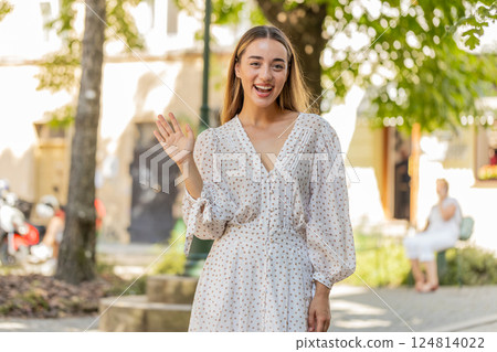 Woman in dress smiling friendly at camera waving hands gesturing hi greeting goodbye on city street Woman in dress smiling friendly at camera waving hands gesturing hi greeting goodbye on city street 124814022
