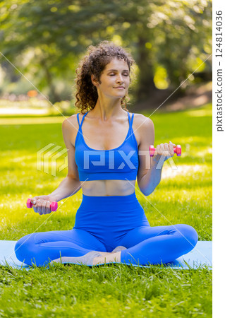 Sportive woman exercising weightlifting with dumbbells sitting on yoga mat in park on sunny day Sportive woman exercising weightlifting with dumbbells sitting on yoga mat in park on sunny day 124814036