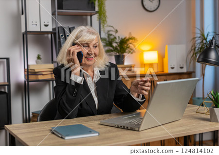 Senior Caucasian businesswoman operator using smartphone and laptop computer at home office desk Senior Caucasian businesswoman operator using smartphone and laptop computer at home office desk 124814102