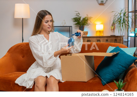 Dissatisfied annoyed young woman opening cardboard box receive broken wireless headphones at home Dissatisfied annoyed young woman opening cardboard box receive broken wireless headphones at home 124814165