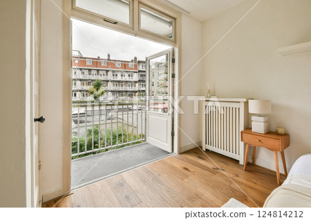 Bright and inviting bedroom featuring an open door that leads to a charming balcony, showcasing an urban landscape with greenery and lovely architecture. 124814212