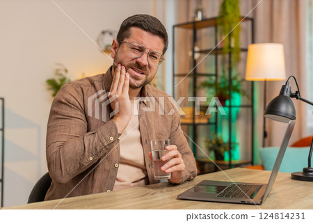 Man freelancer sitting at home office drinking cold water feeling toothache touching mouth at home 124814231