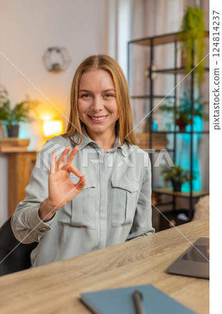 Happy Caucasian woman looking approvingly at camera showing OK gesture sitting at home office table Happy Caucasian woman looking approvingly at camera showing OK gesture sitting at home office table 124814237