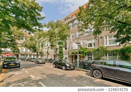 A picturesque view of a residential street lined with trees and parked cars. The buildings feature a blend of modern and traditional architecture, enhancing the local charm. 124814270