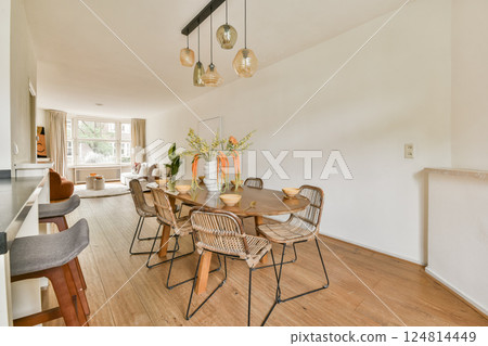 A bright and modern dining area featuring natural wood flooring, elegant lighting fixtures, and stylish rattan chairs complemented by floral decor. 124814449