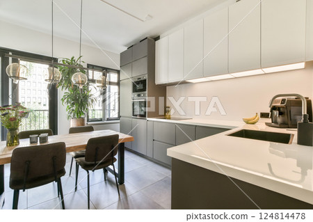 A sleek modern kitchen featuring minimalist cabinetry and stylish furniture, illuminated by natural light from large windows, creating a welcoming atmosphere. 124814478