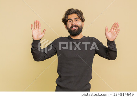 Handsome young Indian man with long sleeves waving hello with his palm against beige background Handsome young Indian man with long sleeves waving hello with his palm against beige background 124814649