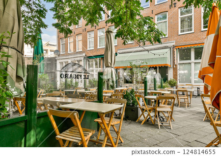 A quaint outdoor cafe scene features wooden tables and chairs nestled among lush greenery, with colorful umbrellas adding a vibrant touch to the setting. 124814655