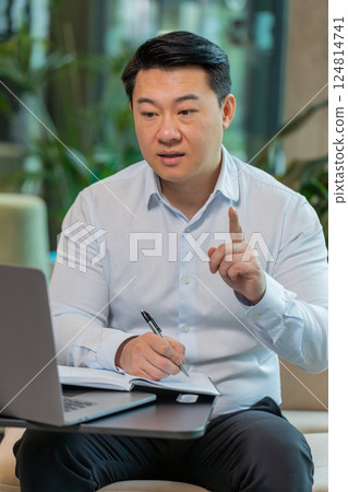 Chinese businessman writing down notes while attending online video class in modern office lobby 124814741