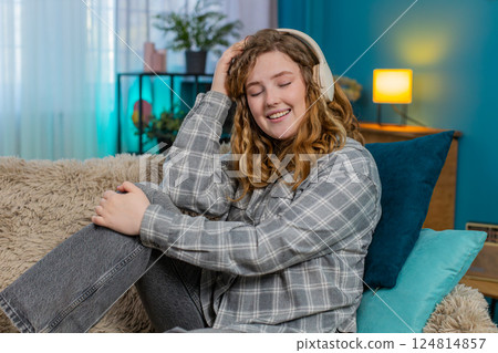 Woman sitting on sofa wearing headphones dancing with closed eyes enjoying favorite relaxing music 124814857