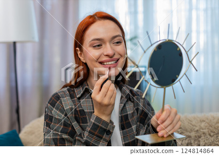 Young pretty woman applying makeup looking in mirror smiling while drawing lips with lipstick gently 124814864