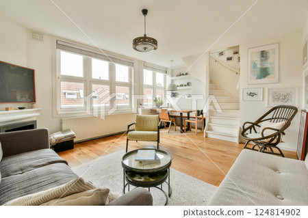 A bright and inviting modern living room featuring stylish furniture, large windows, and an elegant staircase, perfect for relaxation and gatherings. 124814902