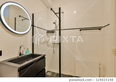 Elegant bathroom featuring a minimalist design, circular mirror, glass shower enclosure, and sleek black cabinetry. Ideal for contemporary home aesthetics. 124815148