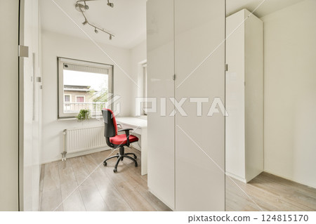 A sleek and modern home office space featuring a white desk, a red chair, and ample natural light, creating a productive and inviting workspace. 124815170