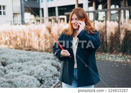 Young adult smiling professional business woman holding smartphone and folder with files while walking in urban city street. Young adult smiling professional business woman holding smartphone and folder with files while walking in urban city street. 124815287