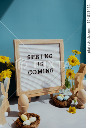 Phrase SPRING IS COMING traditional greeting to christian holiday. Festive mood composition with bunnies and yellow flowers on pastel blue background. Sustainable natural materials Phrase SPRING IS COMING traditional greeting to christian holiday. Festive mood composition with bunnies and yellow flowers on pastel blue background. Sustainable natural materials 124815431