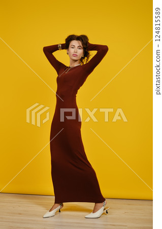 Stylish woman in a long maroon dress poses confidently against a yellow backdrop Stylish woman in a long maroon dress poses confidently against a yellow backdrop 124815589