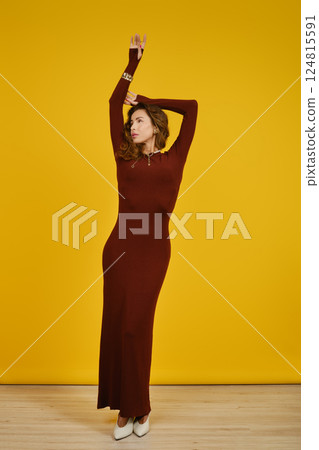 Confident woman in long burgundy dress poses against bright yellow background indoors Confident woman in long burgundy dress poses against bright yellow background indoors 124815591