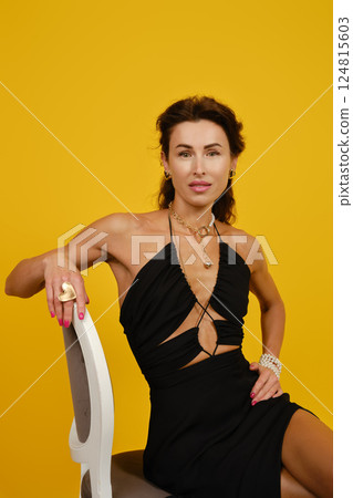 Elegant woman poses confidently in a stylish black dress against a vibrant yellow background Elegant woman poses confidently in a stylish black dress against a vibrant yellow background 124815603