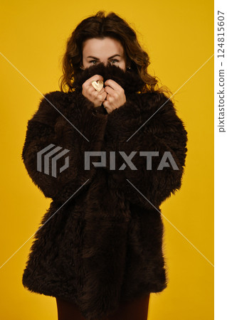Woman in a cozy brown fur coat playfully hiding her face against a bright yellow background 124815607