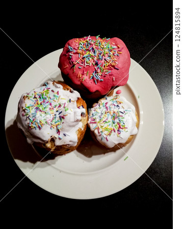 Three easter cakes decorated with white and pink icing, colorful sprinkles scattered, on white plate, dark background Three easter cakes decorated with white and pink icing, colorful sprinkles scattered, on white plate, dark background 124815894