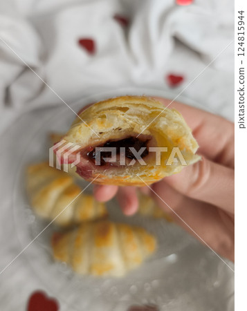 Hand holding warm croissant with jam, resting near heart shaped decor signaling romantic valentine's morning mood 124815944