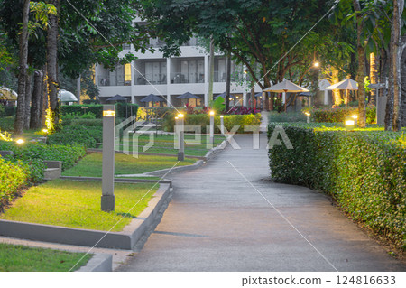 An illuminated asphalt path with lamps under the crowns of green trees in the garden. The backdrop of a hotel in a tropical luxury resort An illuminated asphalt path with lamps under the crowns of green trees in the garden. The backdrop of a hotel in a tropical luxury resort 124816633