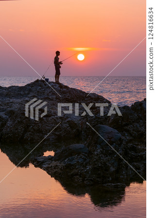 Beautiful seascape at sunset in kaolak thailand - A fisherman's figure on a textured rock by the sea against a colorful purple pink sunset. Sea fishing lifestyle Beautiful seascape at sunset in kaolak thailand - A fisherman's figure on a textured rock by the sea against a colorful purple pink sunset. Sea fishing lifestyle 124816634