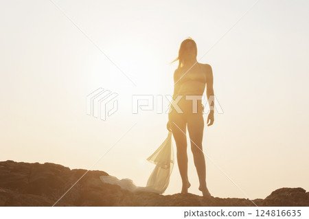 Silhouette of a woman in a swimsuit with a transparent cape in her hands standing on a rock against the sunset, close-up. Silhouette of a woman in a swimsuit with a transparent cape in her hands standing on a rock against the sunset, close-up. 124816635
