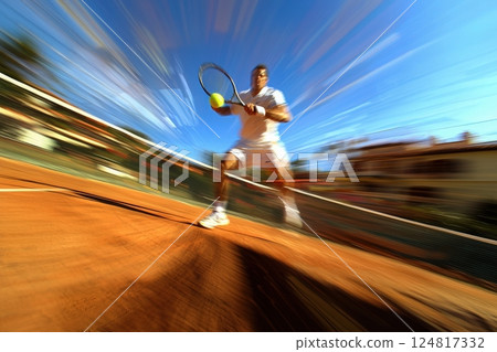 A tennis player in a white outfit strikes a yellow ball with a racket on a clay court. The bright blue sky and blurred background emphasize the players swift motion during the game. 124817332