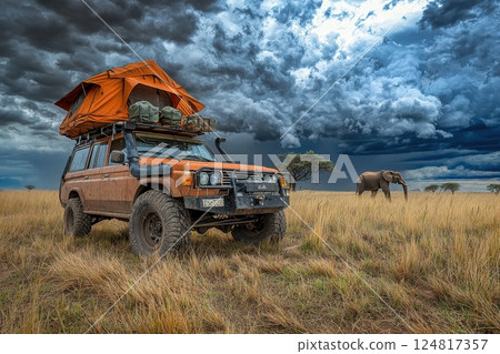 An off-road vehicle equipped with a rooftop tent is parked in a grassy field. An elephant roams nearby under a sky filled with dark, swirling clouds, creating a striking contrast. 124817357