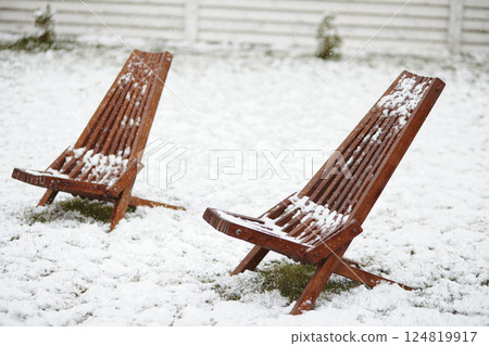 Two wooden outdoor deck chairs stand in a snowy backyard, partially covered in fresh snow. A symbol of unexpected spring frost and sudden cold weather conditions. 124819917