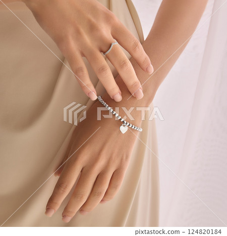 Cropped close-up image of tender female hands wearing silver accessories. Delicate ring and bracelet with heart charm against softly blurred background of light fabric. 124820184