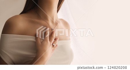 Close-up of elegant woman with engagement golden ring and small diamond on in, wearing silver necklace in heart shape against soft white background Close-up of elegant woman with engagement golden ring and small diamond on in, wearing silver necklace in heart shape against soft white background 124820208