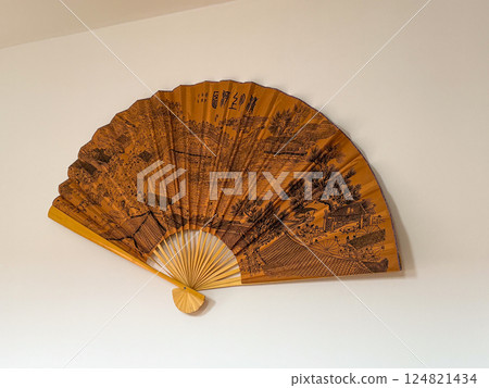 Traditional decorative fan on white wall with detailed historical landscape. Cultural heritage and visual storytelling through Asian artistry. Traditional decorative fan on white wall with detailed historical landscape. Cultural heritage and visual storytelling through Asian artistry. 124821434