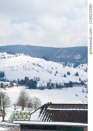 Winter landscape showcasing snow-covered hills, a peaceful farmhouse, and bales of hay resting under a cloudy sky in a serene countryside setting 124822080