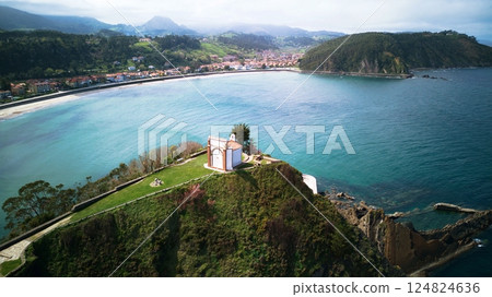 Aerial view of a coastal chapel on a green cliff above turquoise waters, with a seaside town and mountains in the background Aerial view of a coastal chapel on a green cliff above turquoise waters, with a seaside town and mountains in the background 124824636