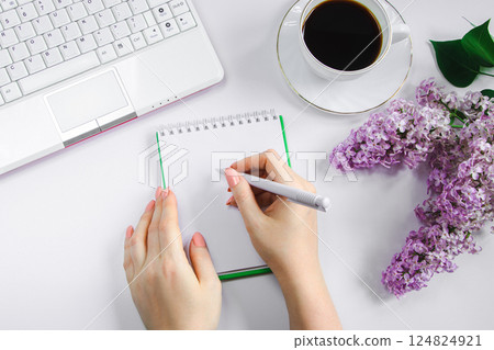 Spring flatlay. Woman works at home on laptop in morning. Cup of coffee and lilac flowers on table. Women's hands write in notepad. 124824921